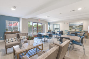 Community Room, 4 four seat tables, coffee table, 4 seats around coffee table, white room, exit to outdoor dining area, landscape painting in the background, attached community kitchen.